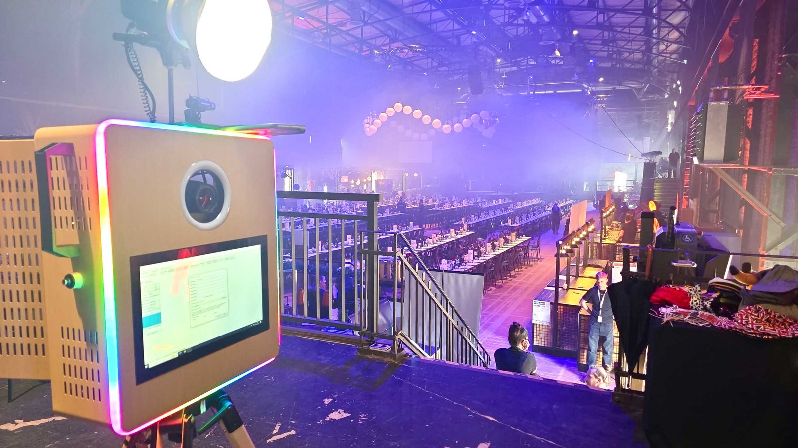 Klassik-Fotobox von Foto Lutz bei der Herweck-Eventparty im E-Werk Saarbrücken — RGB-LED-Box mit Touchscreen im Vordergrund, lange Tafelreihe und blauer Lichtwall der Industriehalle im Hintergrund