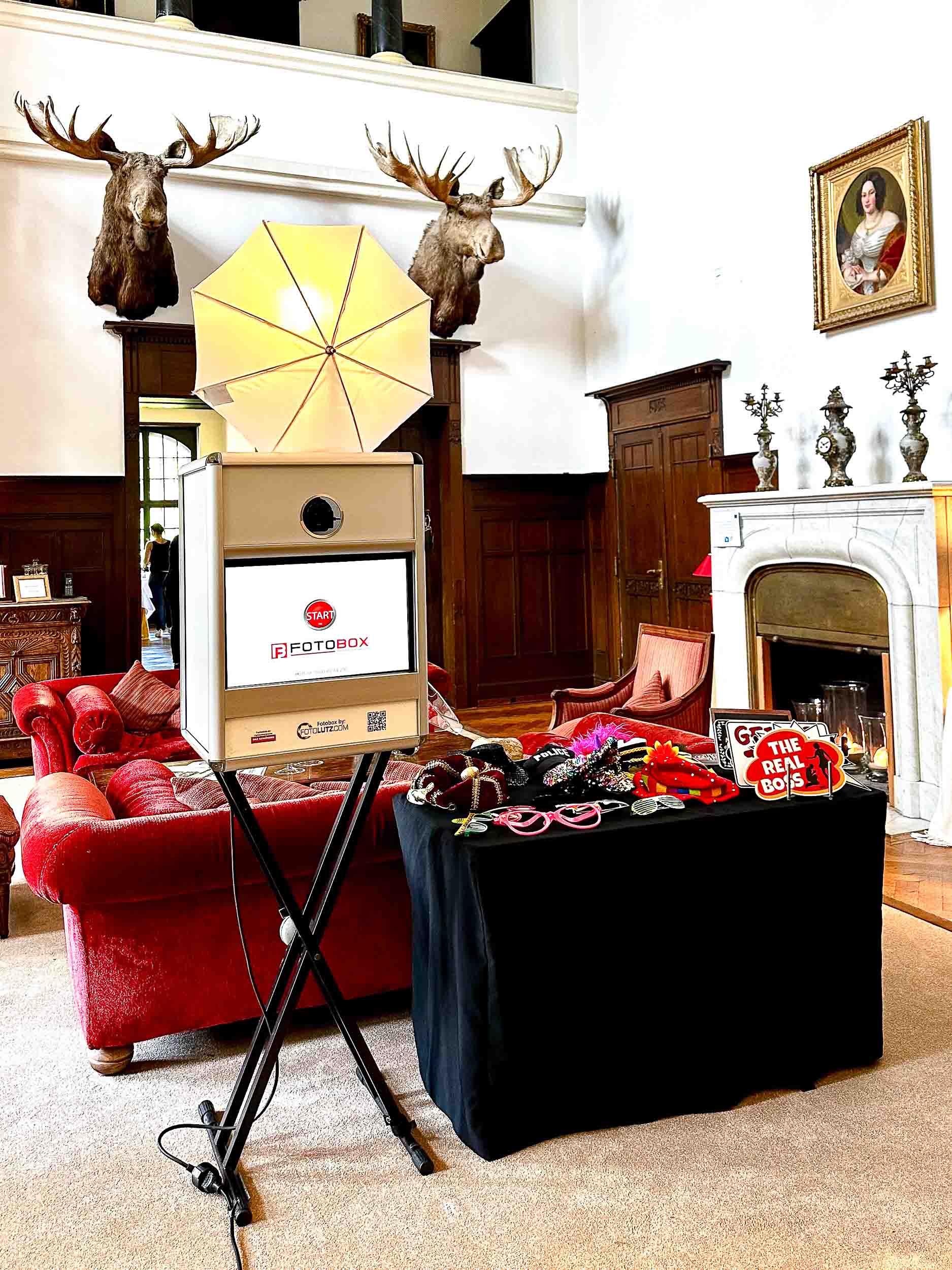 Klassische Fotobox mit Studio-Schirm und Requisitentisch im Einsatz im Jagdschloss-Setting — rote Polstermöbel, Kaminzimmer, antike Elch-Trophäen