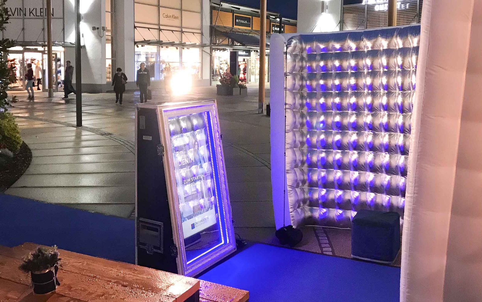 Magic Mirror Outdoor-Setup im Fashion Outlet Center Zweibrücken bei Nacht — blau beleuchteter Spiegel, LED-Cube-Backdrop, Calvin Klein Store im Hintergrund