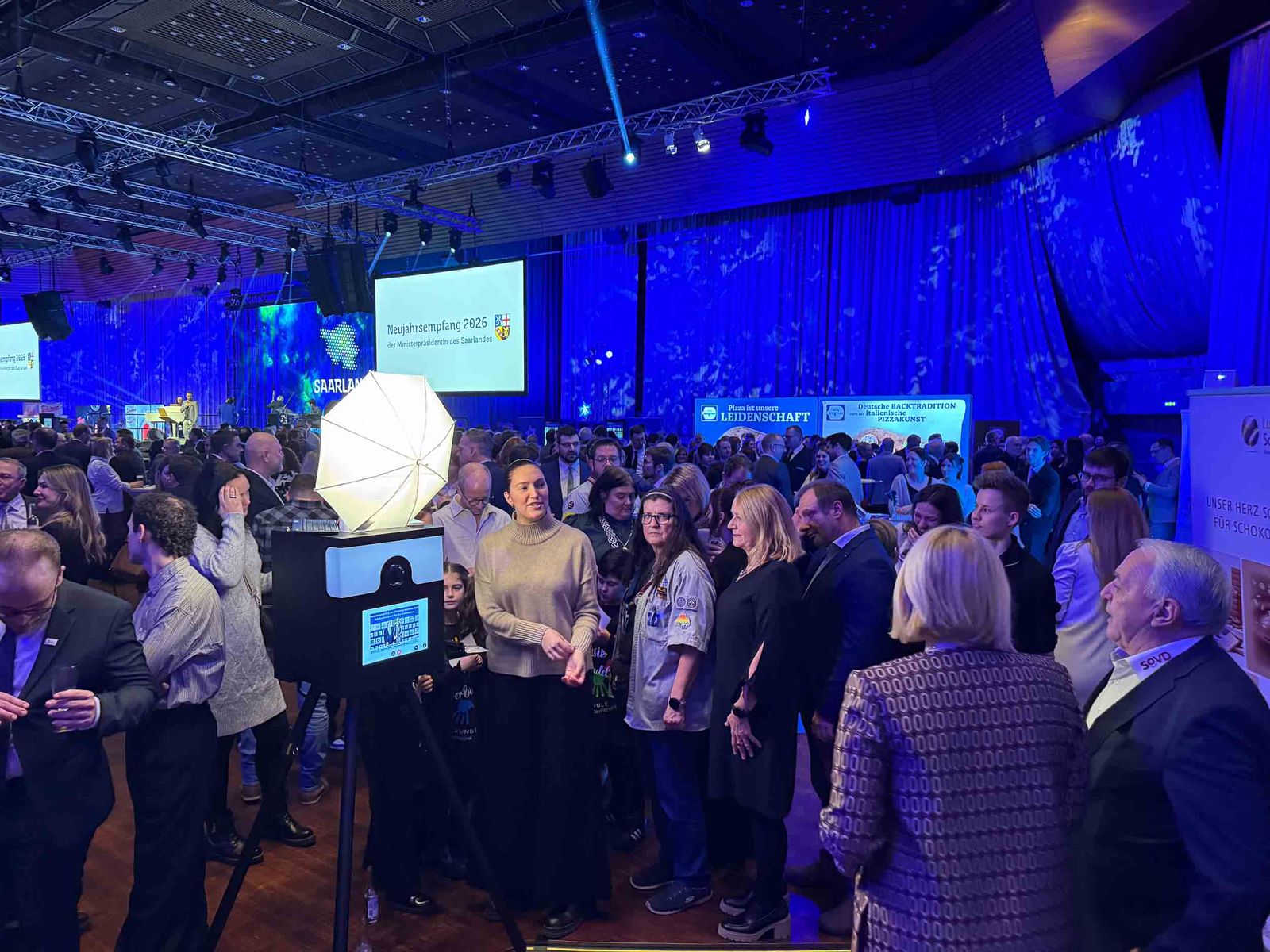 Klassik-Fotobox von Foto Lutz im Einsatz beim Neujahrsempfang 2026 der Ministerpräsidentin in der Saarlandhalle — Studio-Schirm im Vordergrund, Gästegruppe vor blauer Festkulisse mit Bühne und Eventbranding