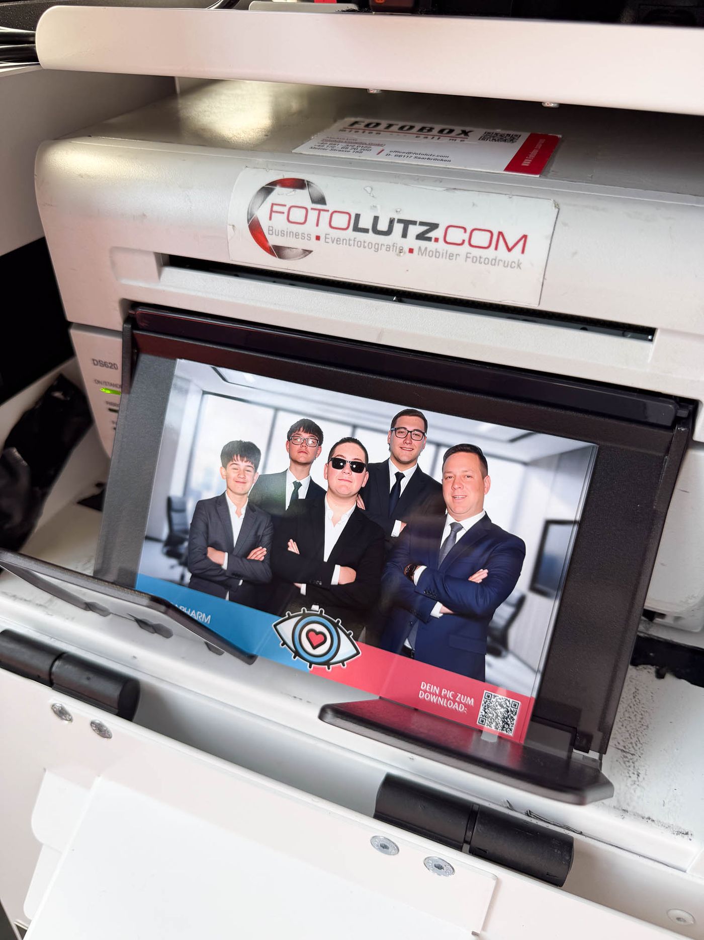 Frischer KI-Sofortdruck im Business-Setting kommt aus dem Foto-Lutz-Drucker — fünf Männer als Business-Gruppe vor Conference-Room mit Skyline, Ursapharm-Branding und QR-Code, Foto-Lutz Branding auf dem Drucker sichtbar