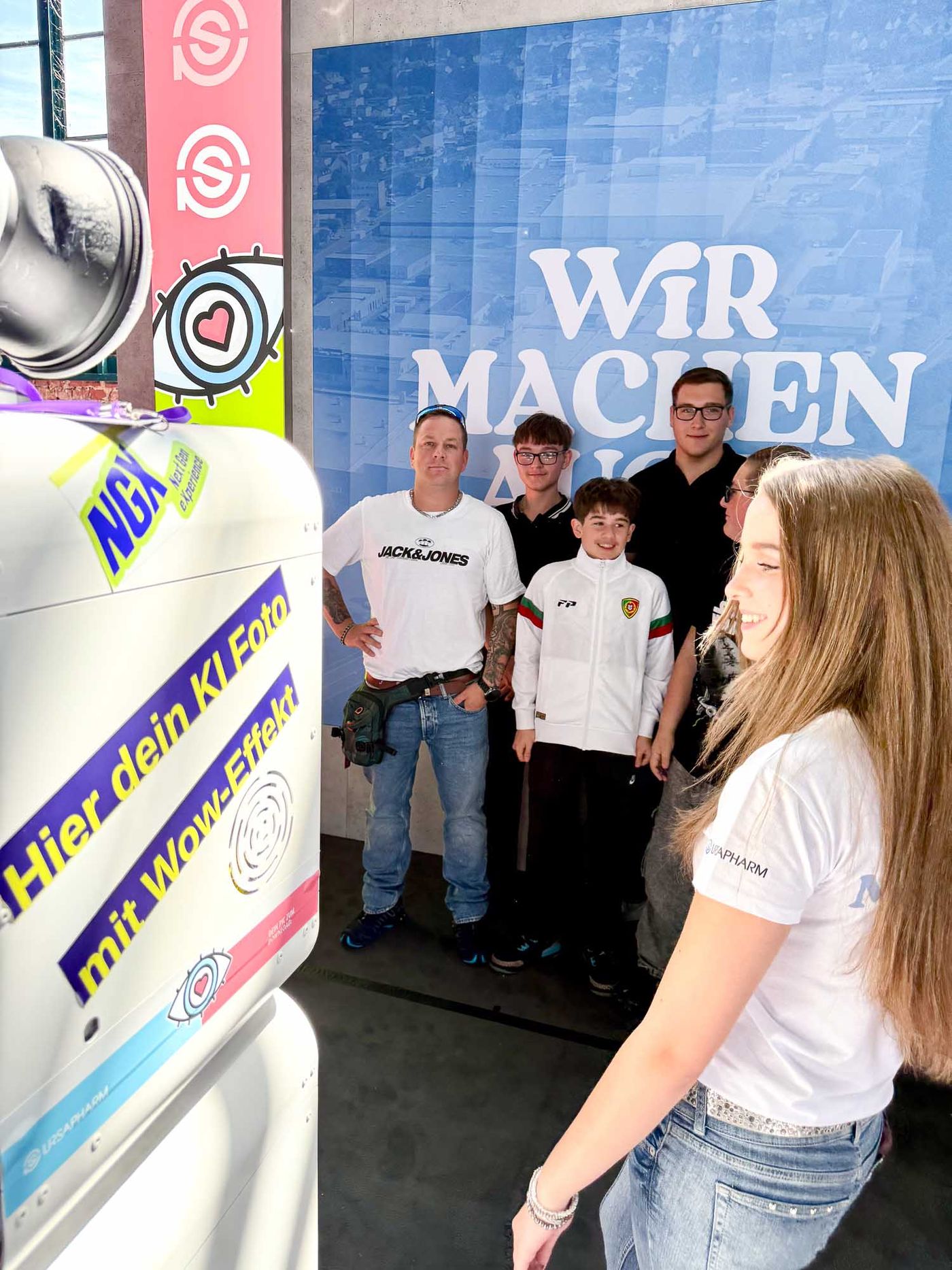 Freundesgruppe posiert vor dem WIR MACHEN AUGE Backdrop am Ursapharm-Stand bei der NextGen eXperience — junge Gäste vor der Box mit Display Hier dein KI-Foto mit Wow-Effekt im Vordergrund