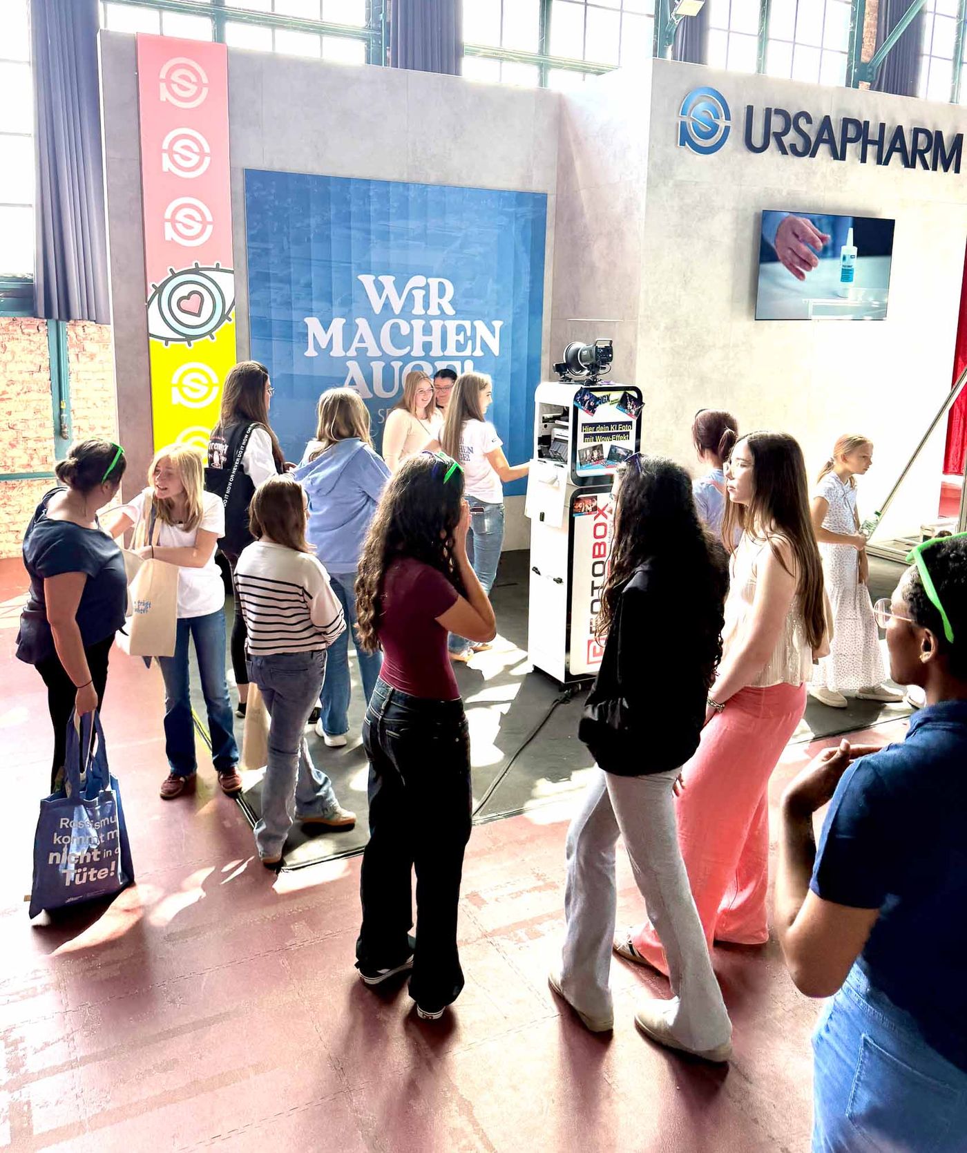 Ursapharm-Messestand WIR MACHEN AUGE bei der NextGen eXperience im E-Werk Saarbrücken — Gästegruppe vor der Foto-Lutz KI-Fotobox, im Industriehallen-Setting