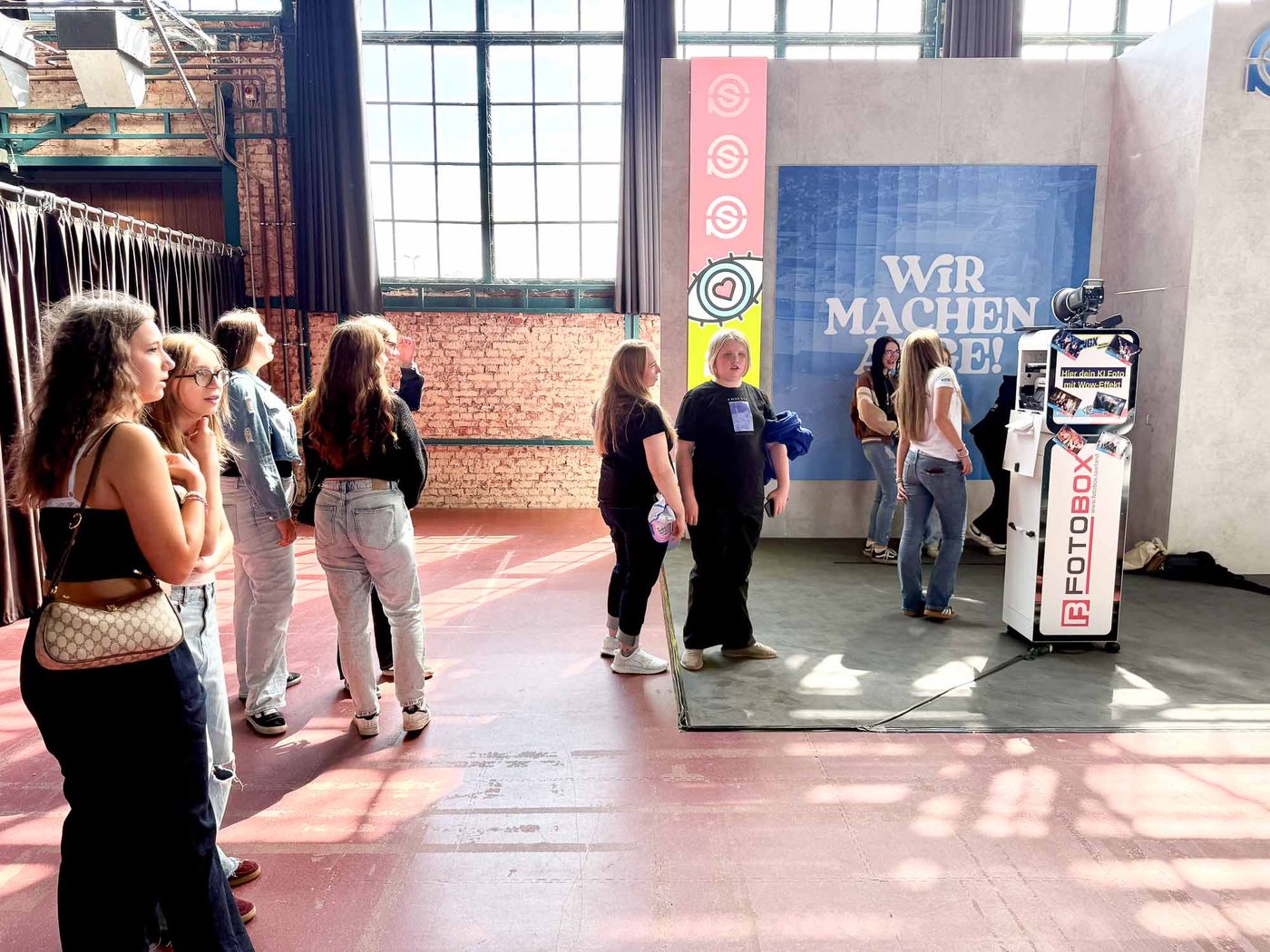 Warteschlange vor der Foto-Lutz KI-Fotobox am Ursapharm-Stand NextGen eXperience — junge Frauen warten bis sie an der Reihe sind, WIR MACHEN AUGE Backdrop im Hintergrund