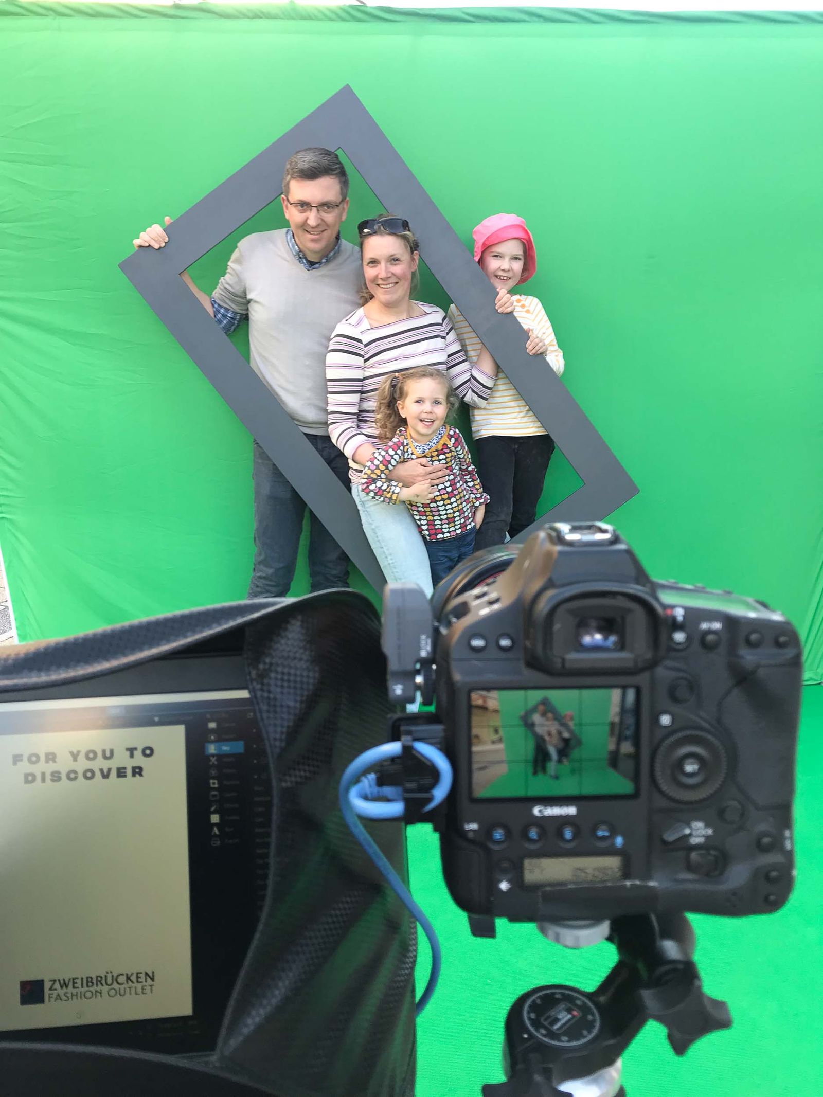 Making-of: Familie posiert mit grauem Frame vor Greenscreen im Fashion Outlet Zweibrücken, Canon EOS-1D Profi-Kamera mit Live-Vorschau im Vordergrund, Computer mit Fashion-Outlet-Druckmotiv