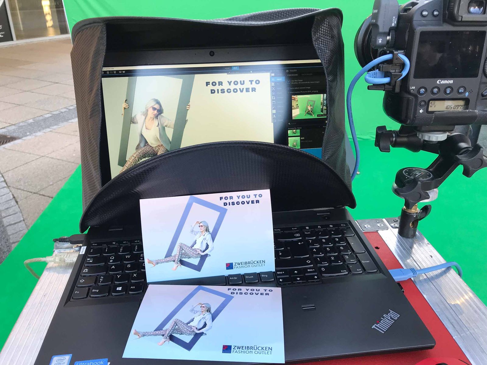 Foto Lutz Pipeline-Setup: Laptop mit Druckvorschau, frischer Sofortdruck FOR YOU TO DISCOVER mit Zweibrücken-Fashion-Outlet-Branding, Canon EOS-1D Kamera im Vordergrund