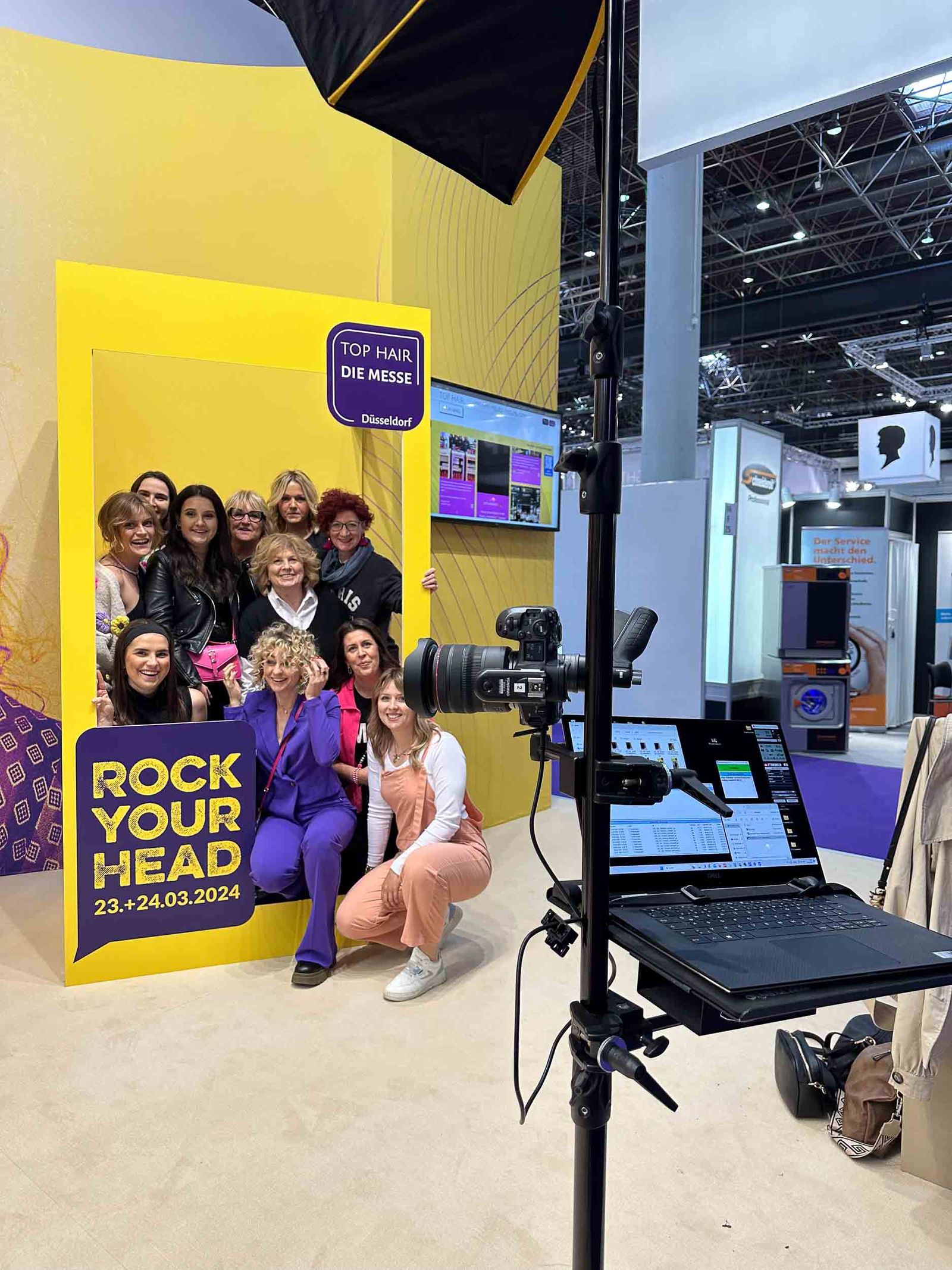 Tophair 2024 Foto-Lutz-Setup — Gruppe Friseur-Frauen posiert im ROCK YOUR HEAD Frame, TOP HAIR Die Messe Düsseldorf Branding, Computer mit Live-Pipeline im Vordergrund